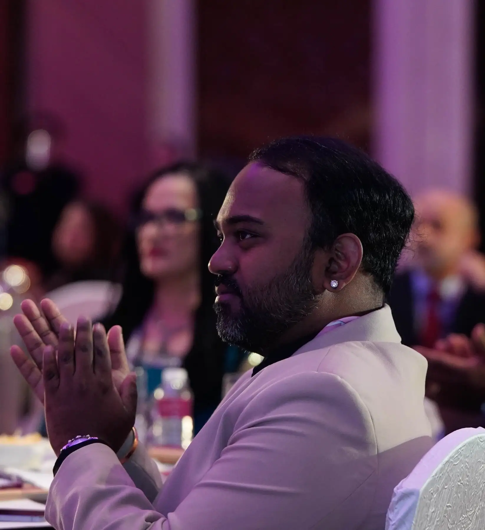 Alex Gomes applauding from a table at the FLUXX Conference, in a pale jacket, seen in profile