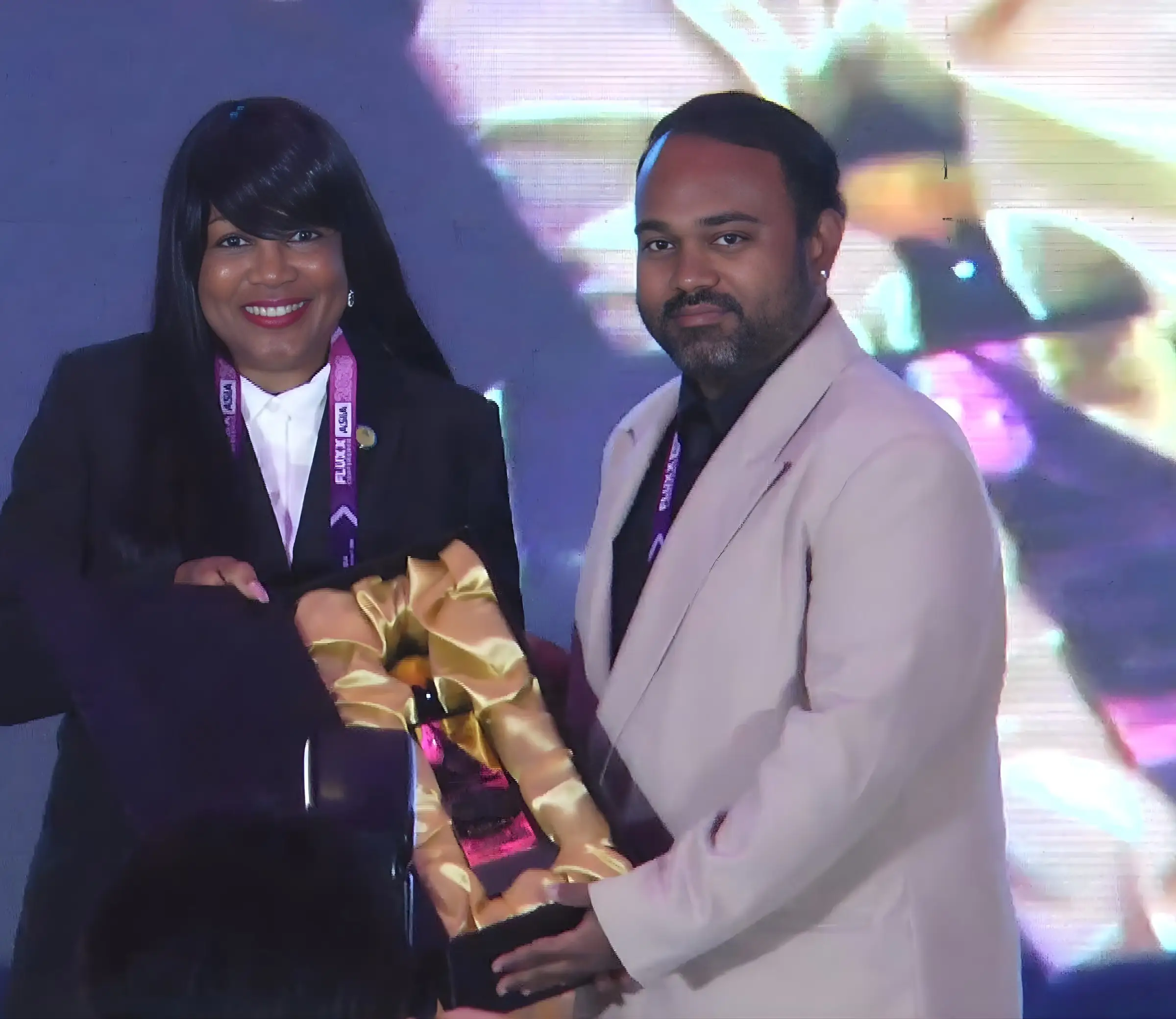 Alex Gomes accepting the award trophy on stage at the FLUXX Conference, presented by a woman in a dark suit